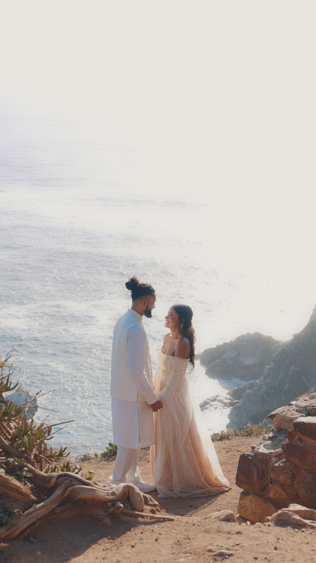 A couple in wedding attire sits on a cliff overlooking a dramatic ocean vista at sunset.