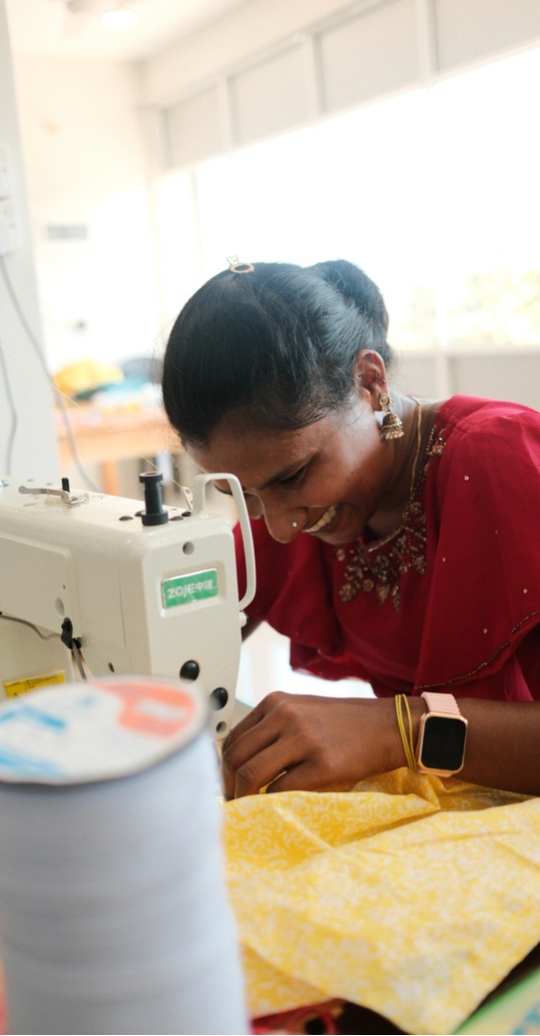 A seamstress in a red top concentrates while working at a sewing machine with yellow fabric.