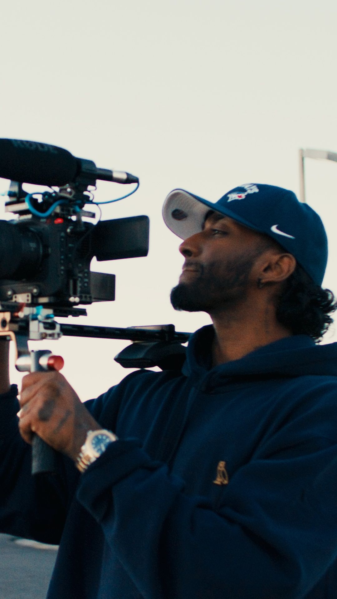 Camera operator in a navy blue hoodie and cap looking through professional video camera lens.