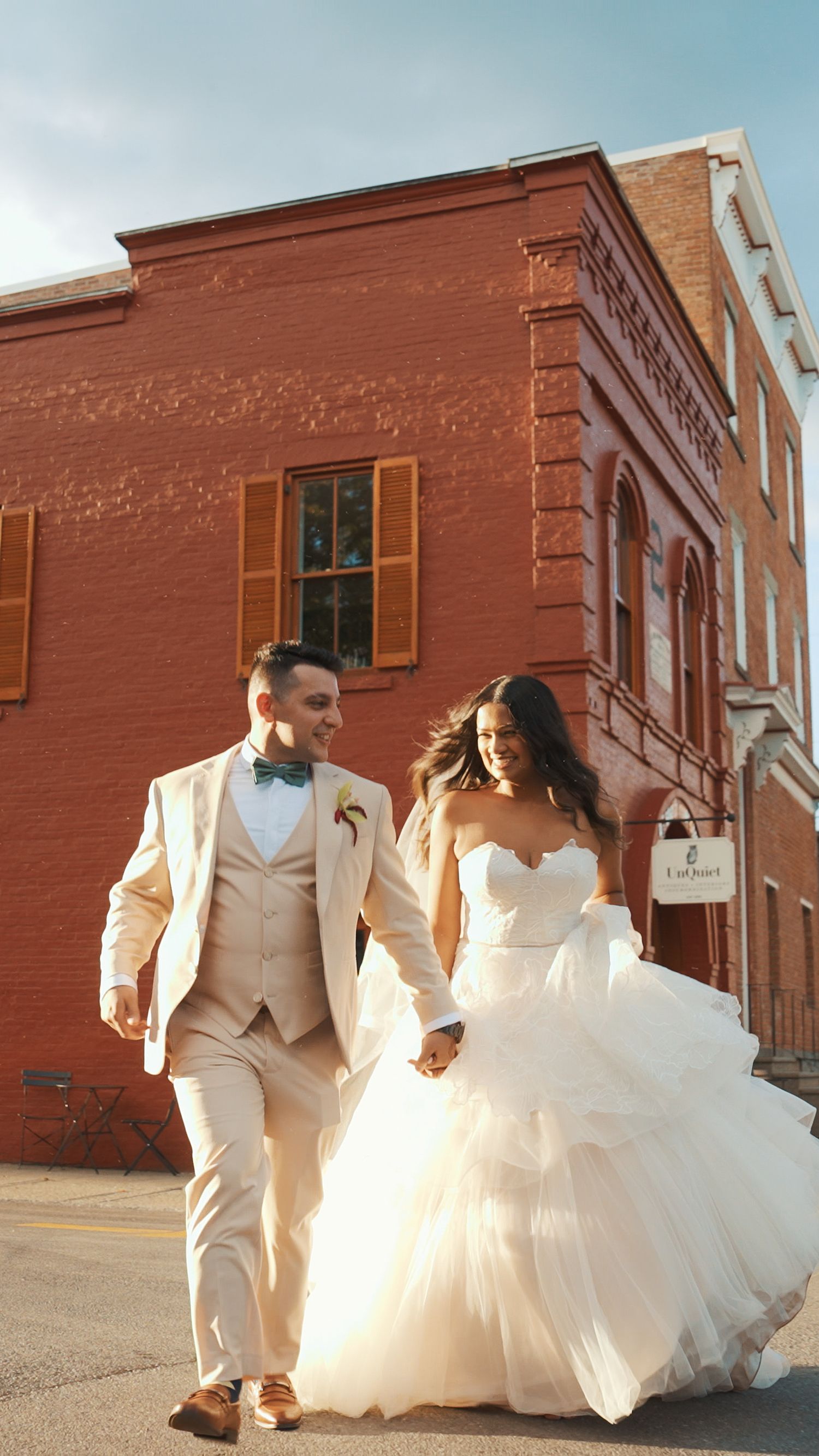 A couple in elegant white wedding attire walks and poses together against a rustic brick building exterior.