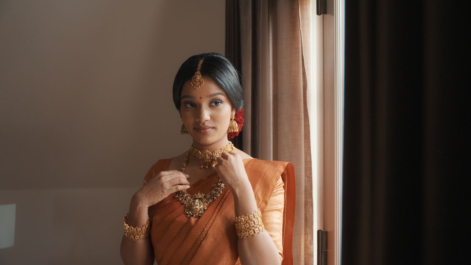 Person wearing elegant orange sari with traditional jewelry stands by window.
