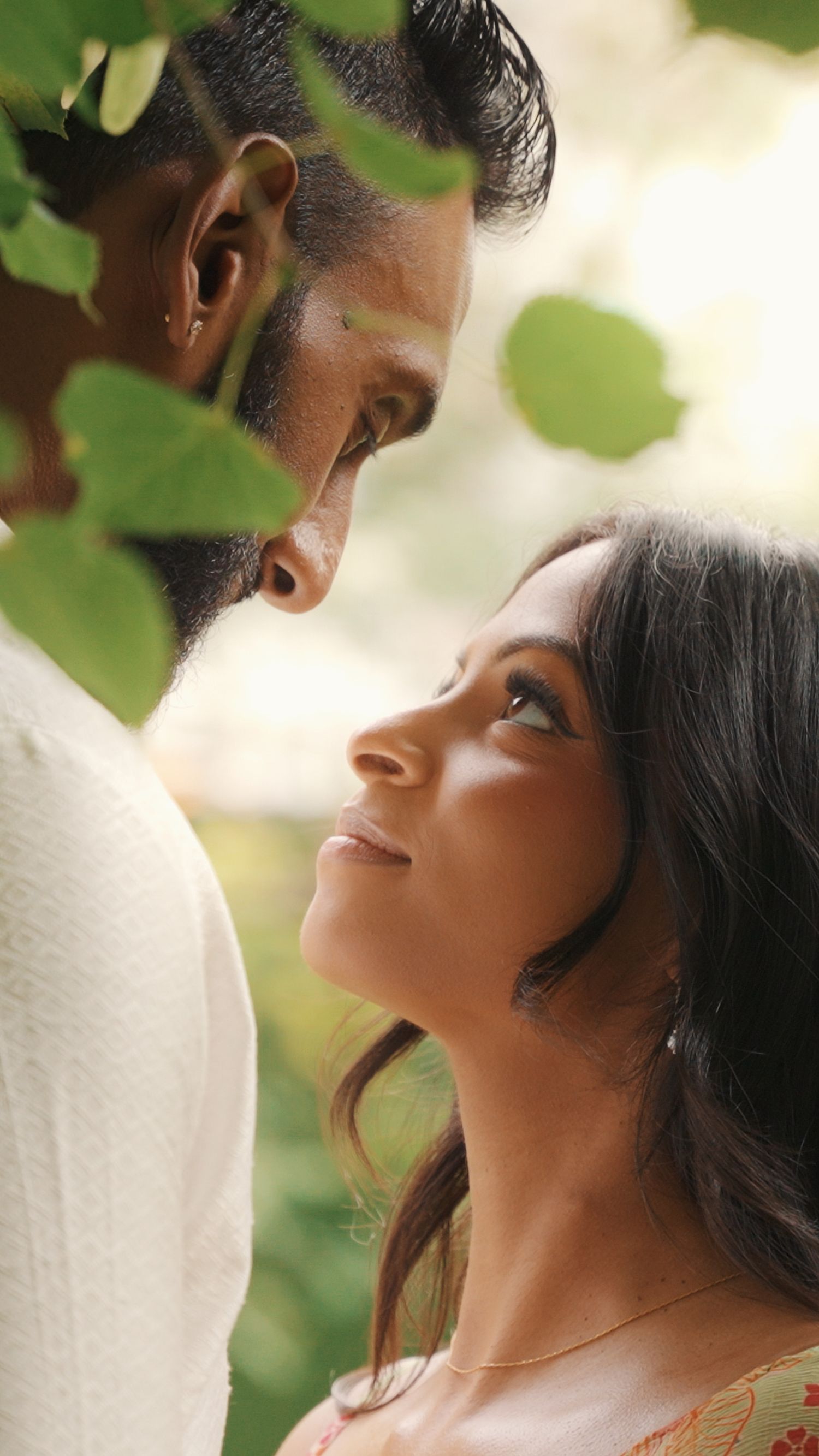 Romantic couple shares an intimate moment under green foliage in soft lighting.