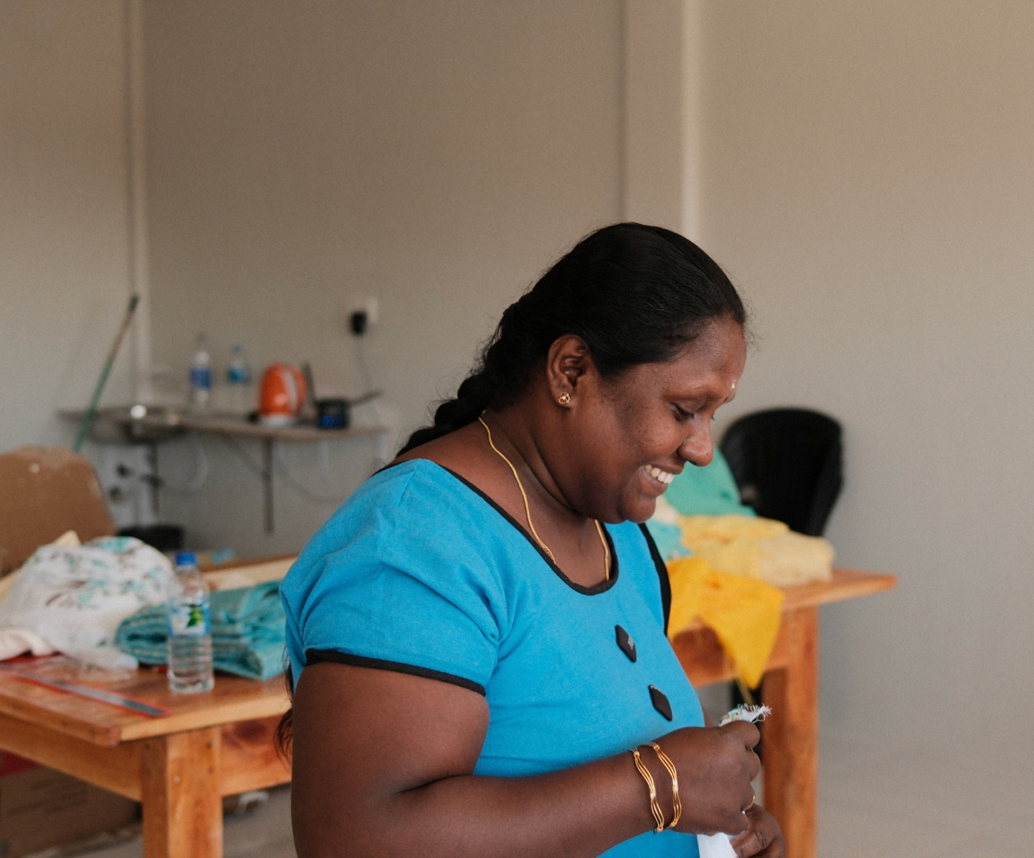 Person in turquoise shirt working at a sewing machine in a bright workshop setting.