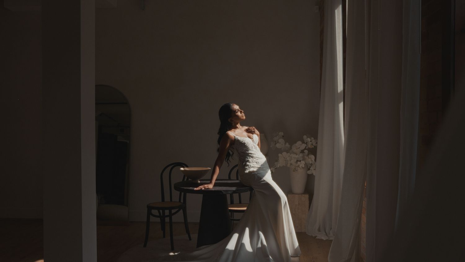 Dancer in white dress poses gracefully near window in moody artistic portrait.