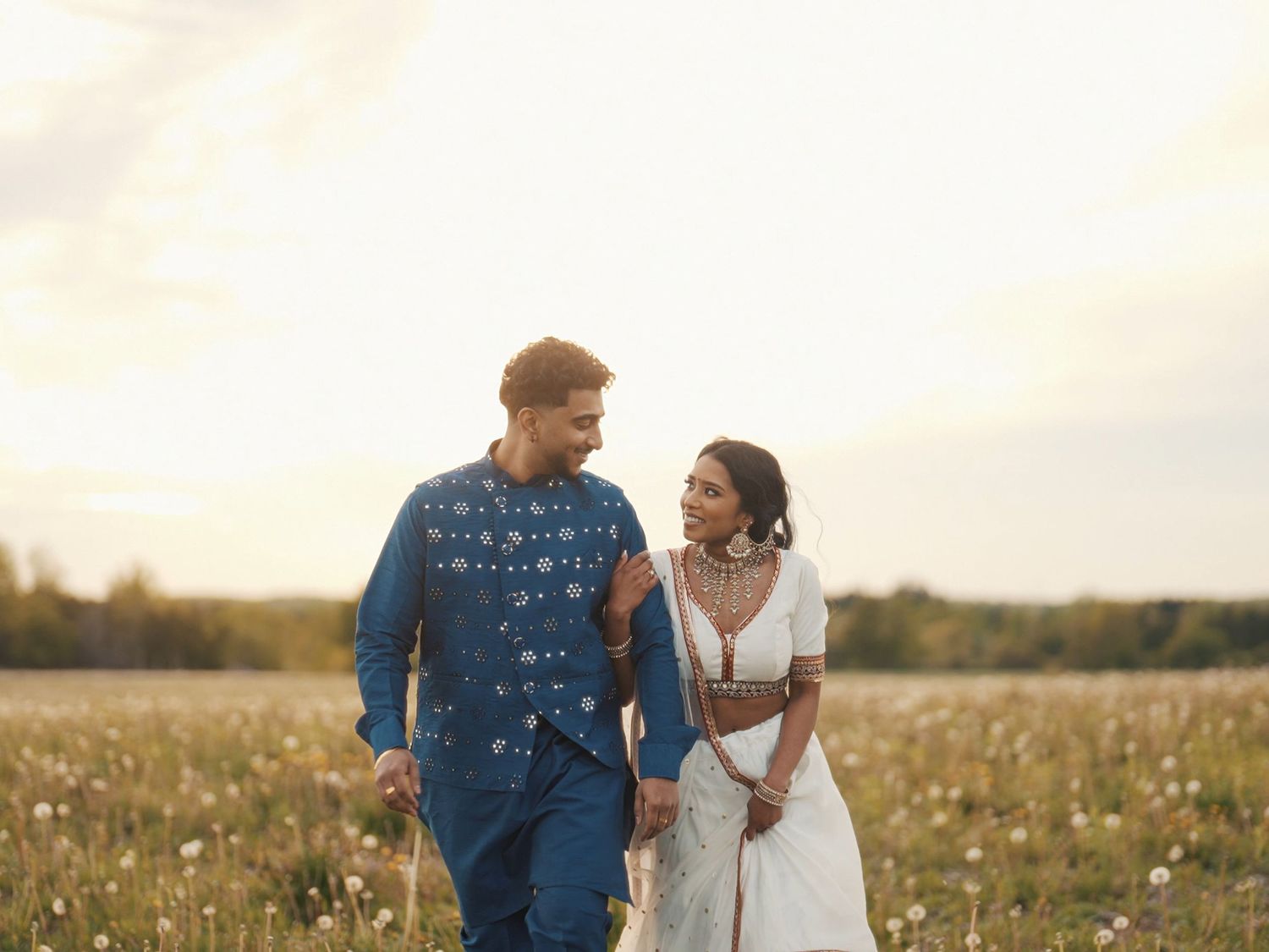 Couple walking hand in hand through a wildflower field at sunset, man in blue kurta, woman in white dress.
