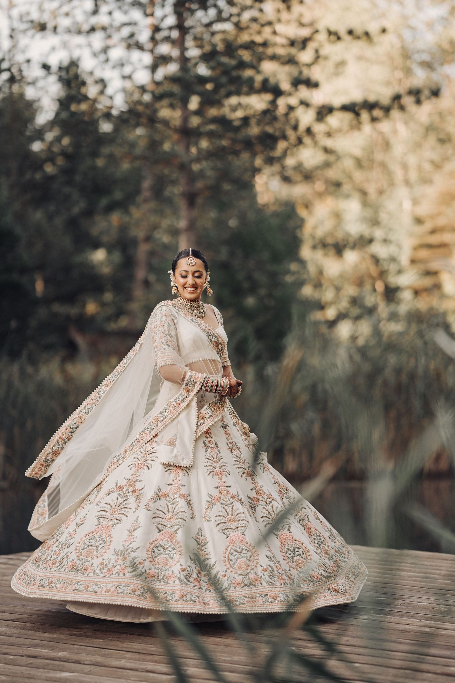An ornate white and gold lehenga twirls elegantly on a wooden deck surrounded by trees.