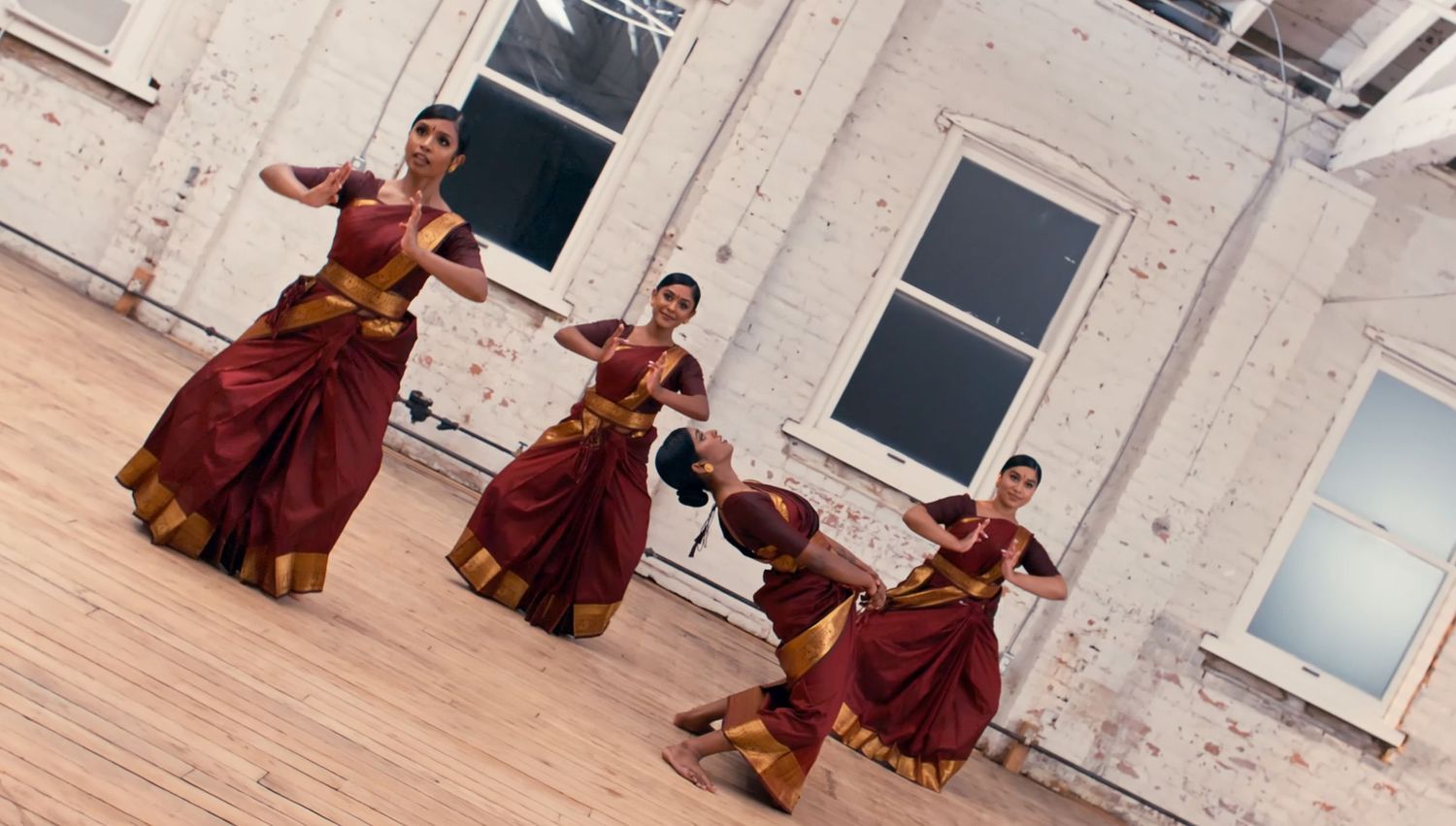 Dancers in flowing brown costumes perform a contemporary dance sequence in a bright studio space.