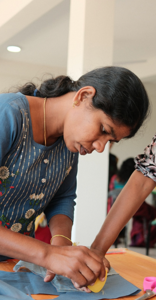 A person in a blue patterned dress leans over a table while carefully writing or drawing on paper.