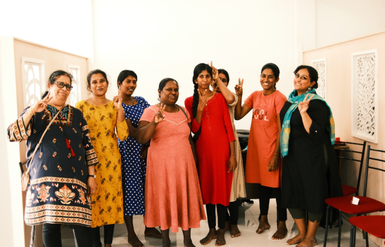 A group of people in colorful outfits stand together against a white wall in a studio-like setting.