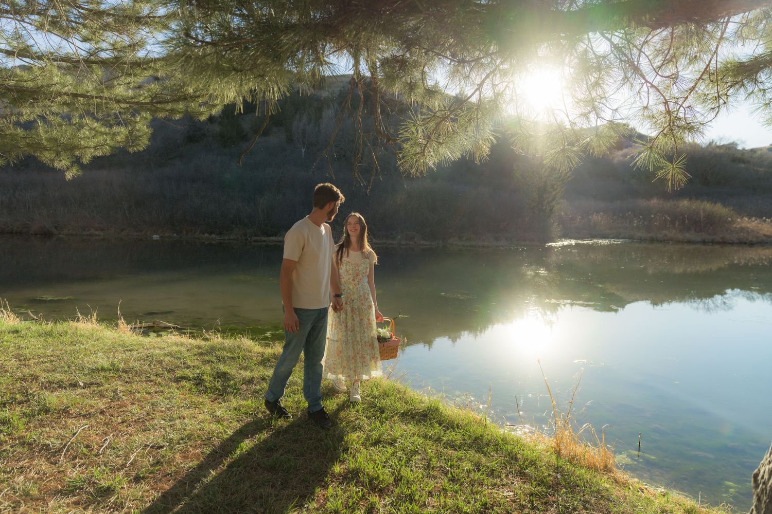 Couple standing by a sunlit pond surrounded by trees and green grass during golden hour.