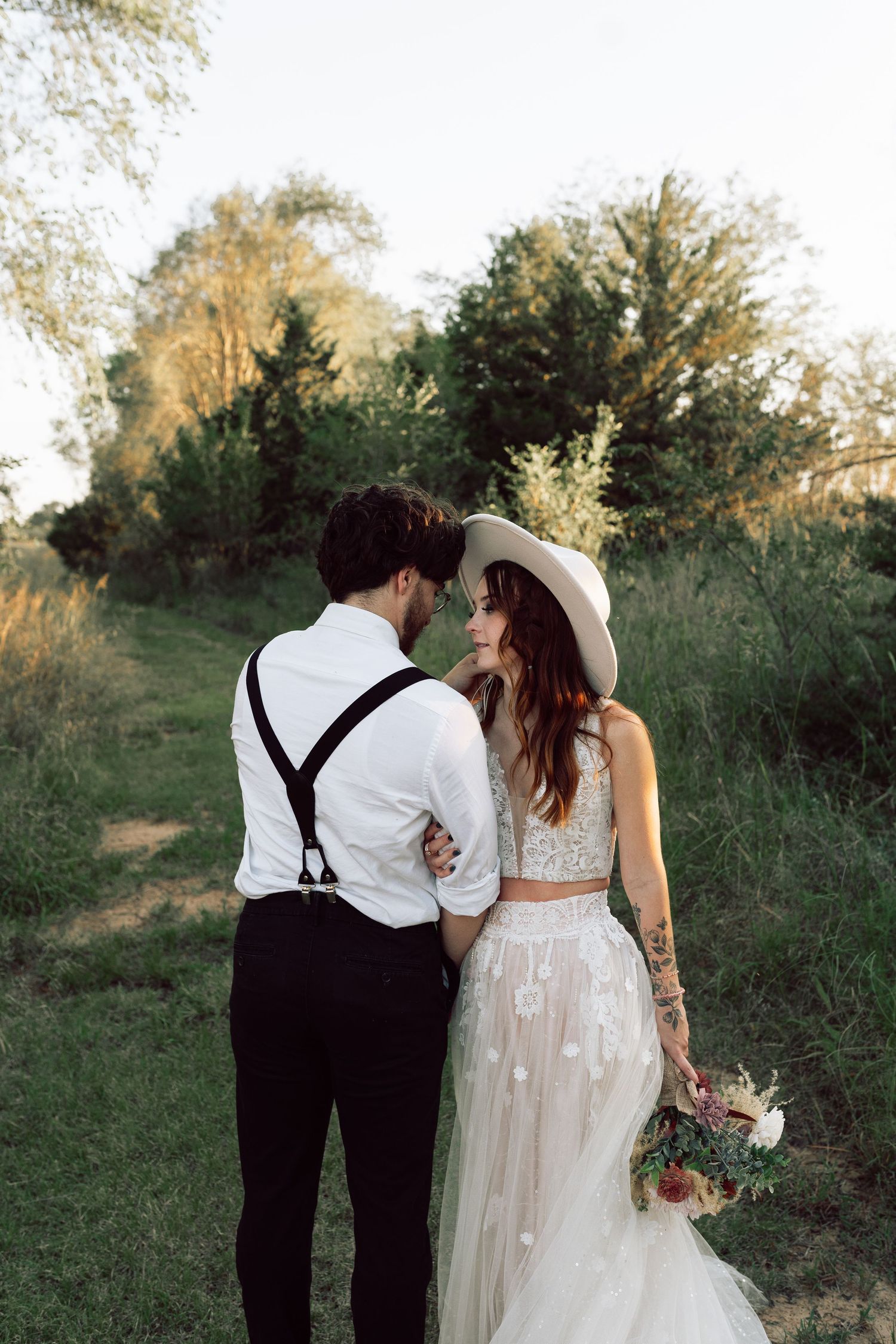 A couple in wedding attire walks hand in hand along a grassy path at sunset.