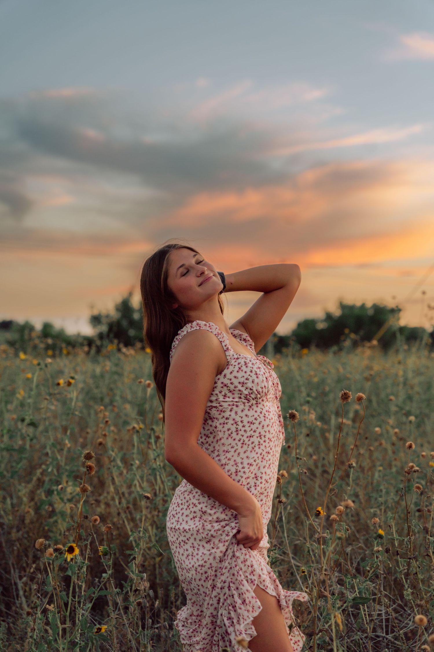 A woman in a floral dress poses gracefully in a wildflower field during golden hour sunset.