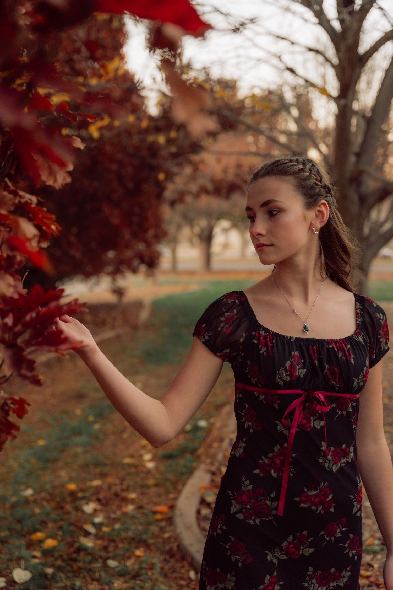 A woman in a dark floral dress touches autumn leaves in a park setting during golden hour.