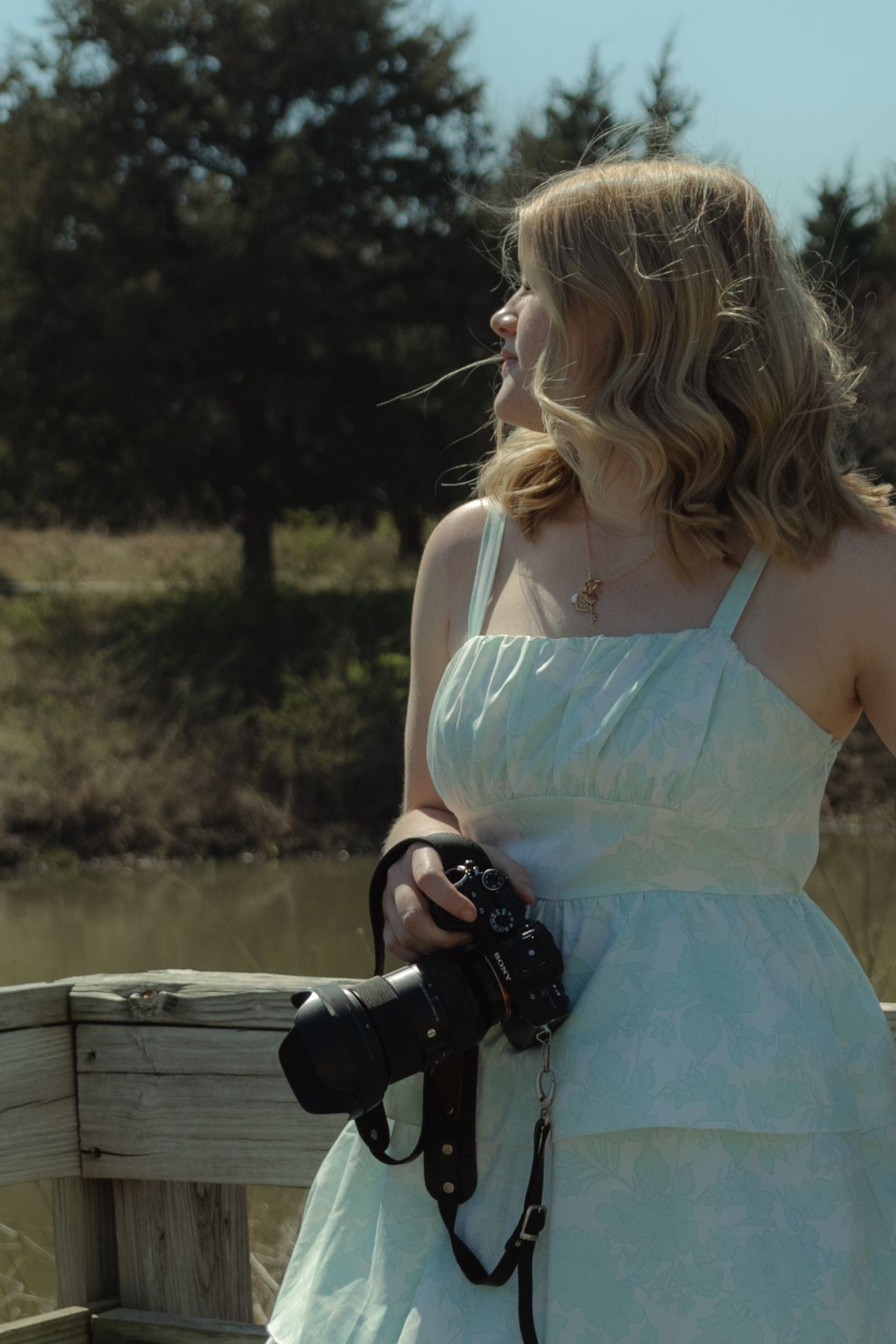 Young blonde woman in light blue sundress holding a camera on a wooden bridge near a pond on a sunny day.