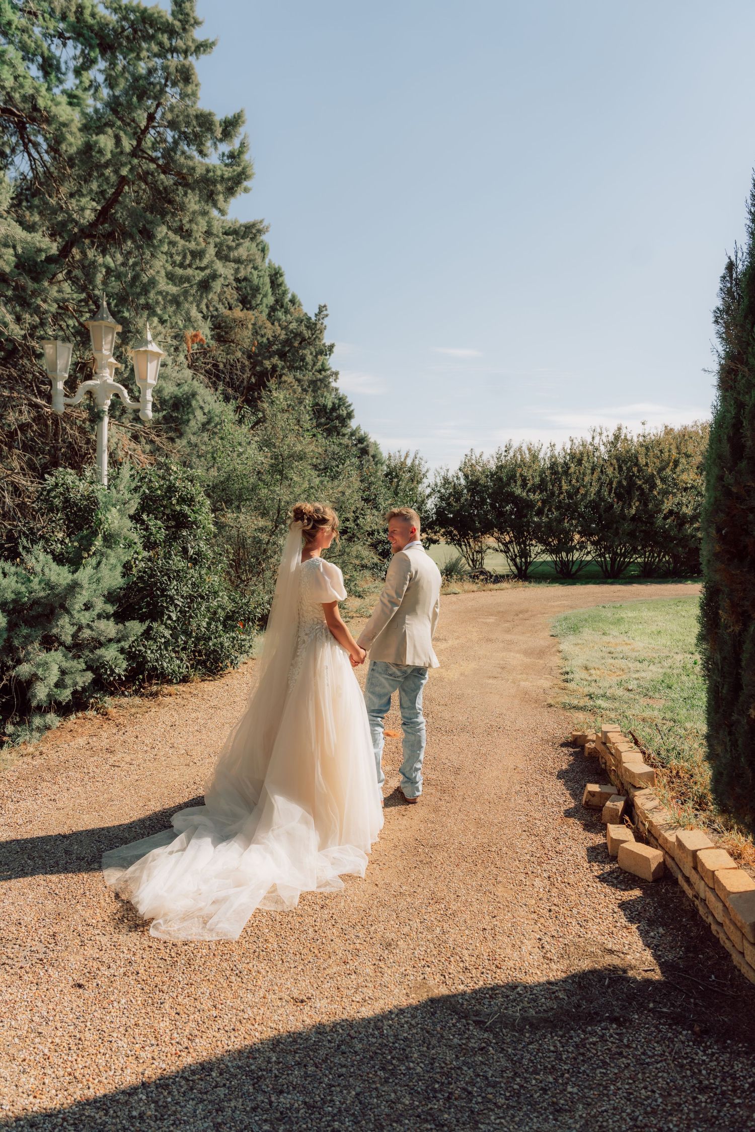 Wedding couple walking along a gravel path surrounded by evergreen trees.