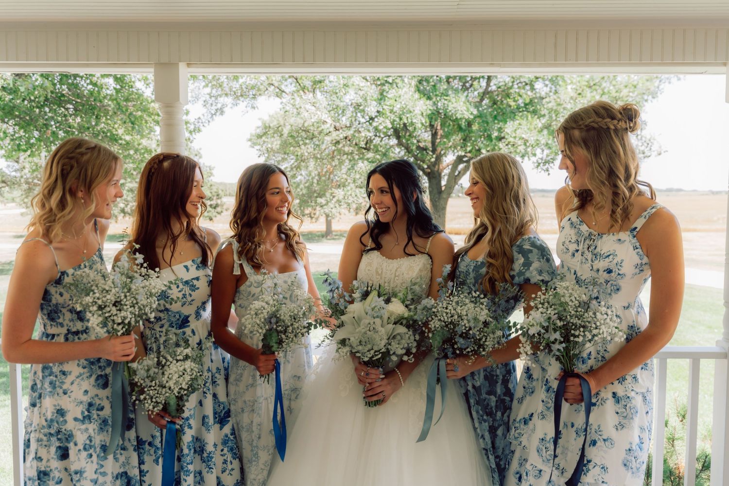 Bridesmaids in blue floral dresses pose together on a covered porch.