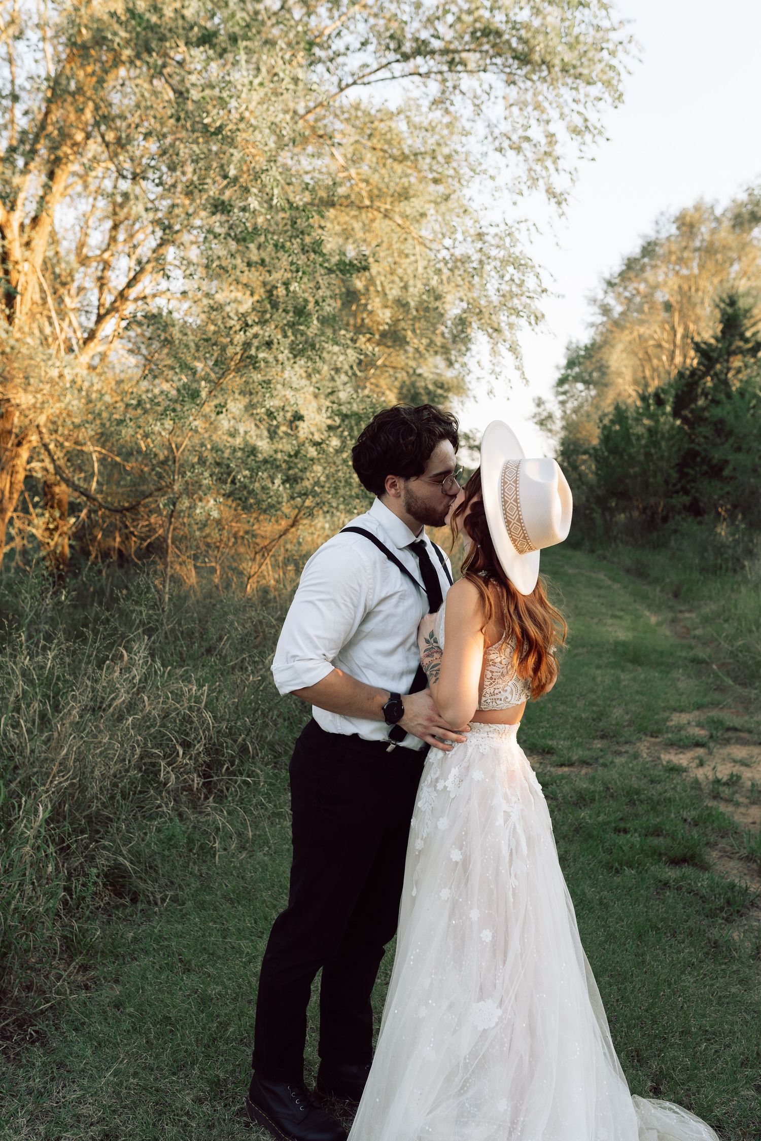A couple in vintage-inspired wedding attire embraces on a nature trail during golden hour.