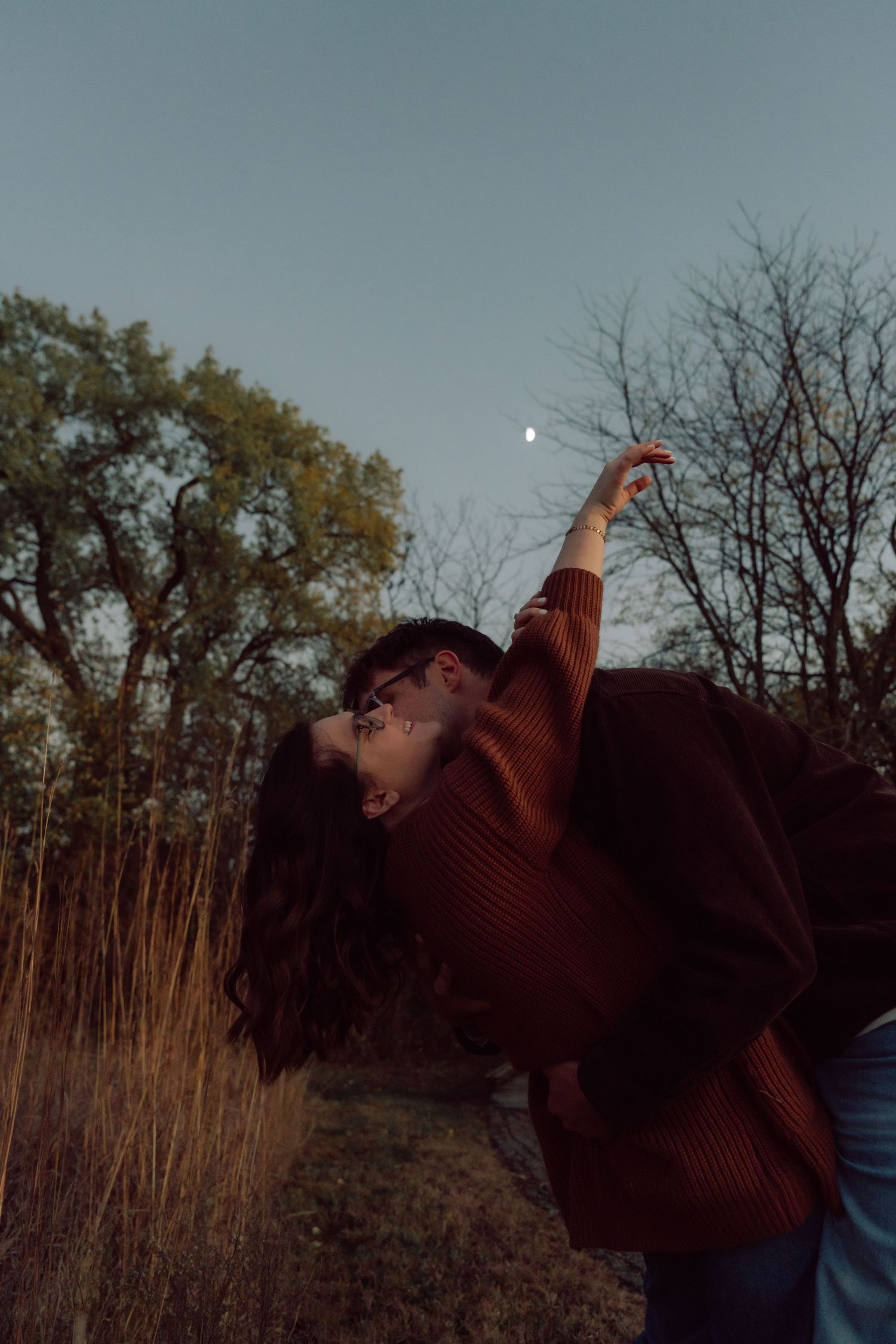 Silhouetted figure in a burgundy sweater reaching up towards the moon in a dusky autumn landscape.