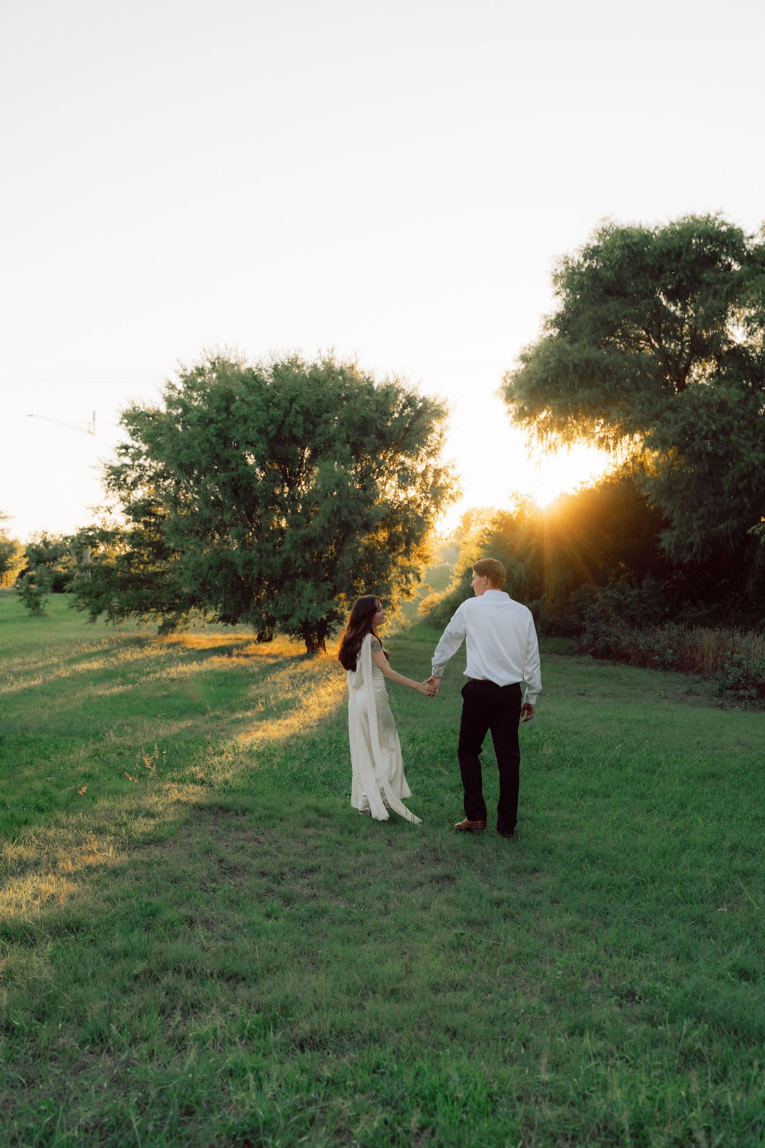 Two people walk hand in hand through a green field at sunset with trees silhouetted against the sky.
