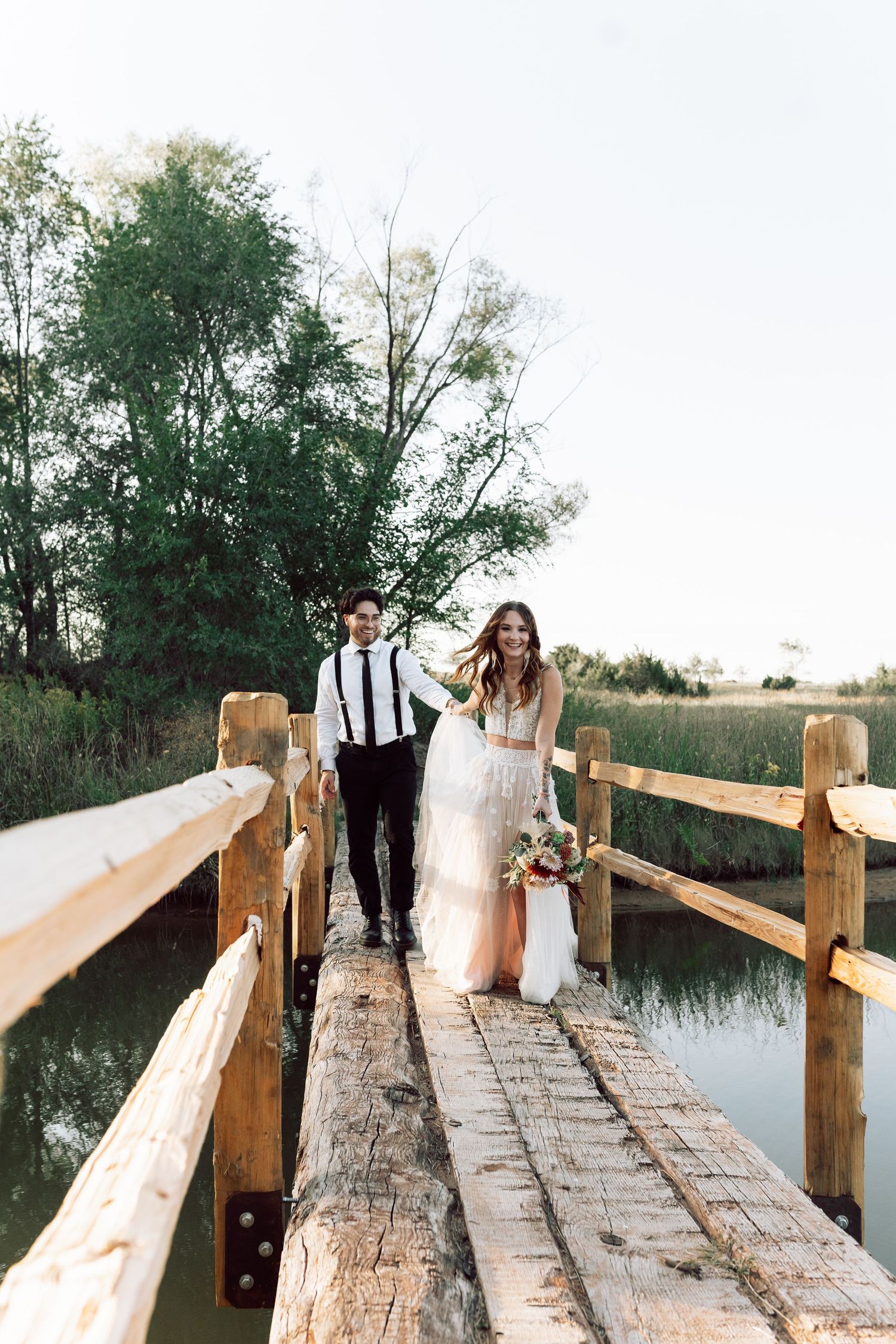 A couple walks along a rustic wooden bridge over a tranquil pond during sunset.