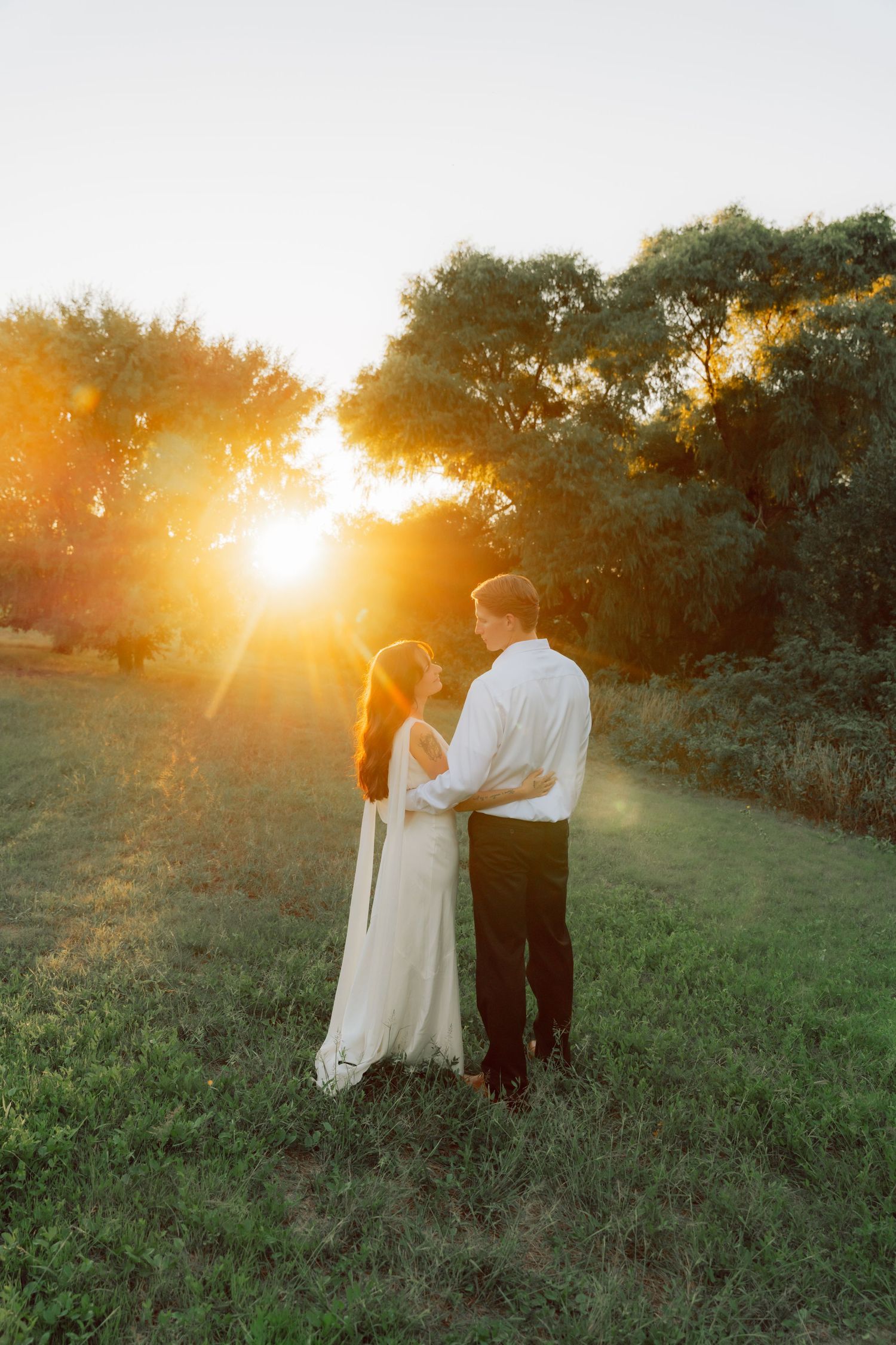 A couple shares an intimate moment in a sunlit field surrounded by nature at dusk.