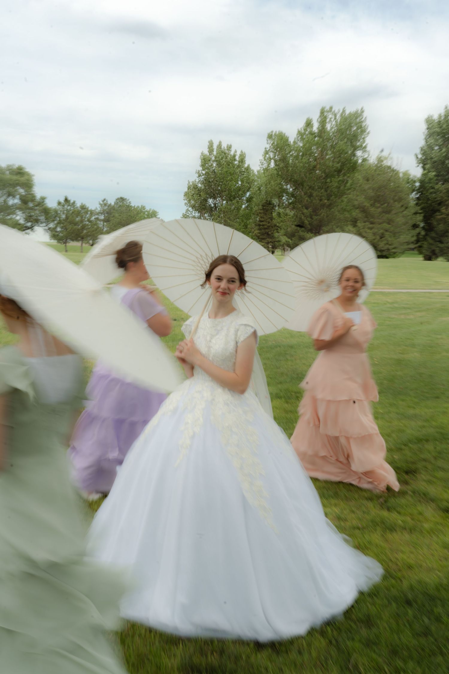 Bride in white ball gown holds parasol with bridesmaids in pastel dresses outdoors on green lawn.