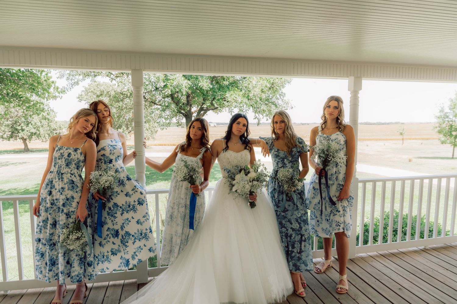 Wedding party in floral blue dresses and white gown gathered on a rustic porch.