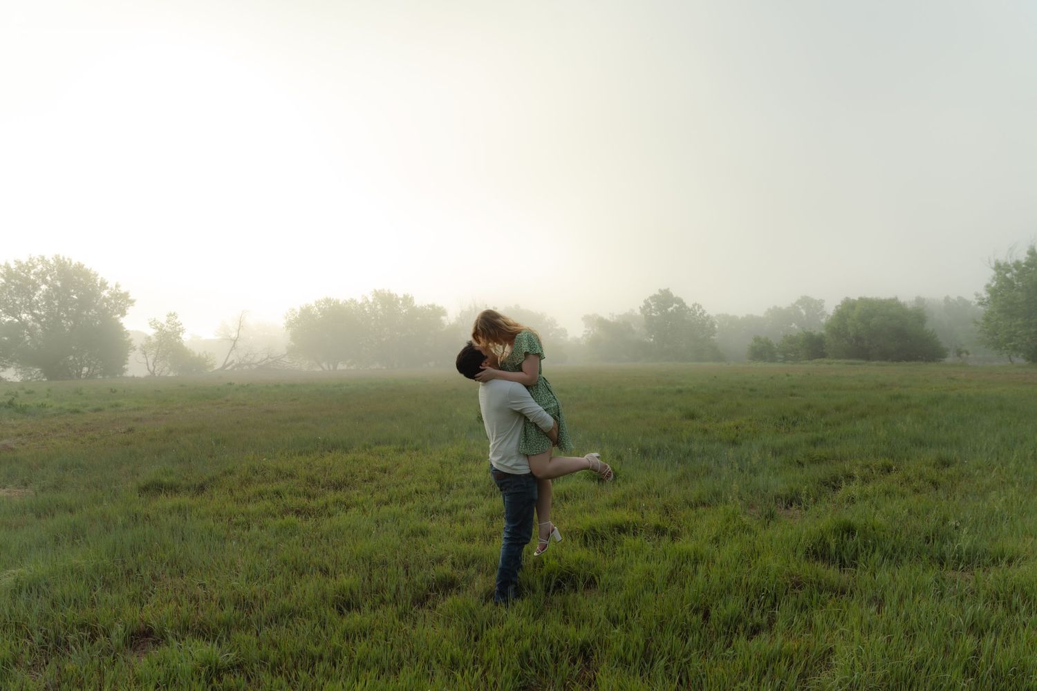 Couple embracing romantically in a misty green field at dawn, surrounded by fog and trees in the background.