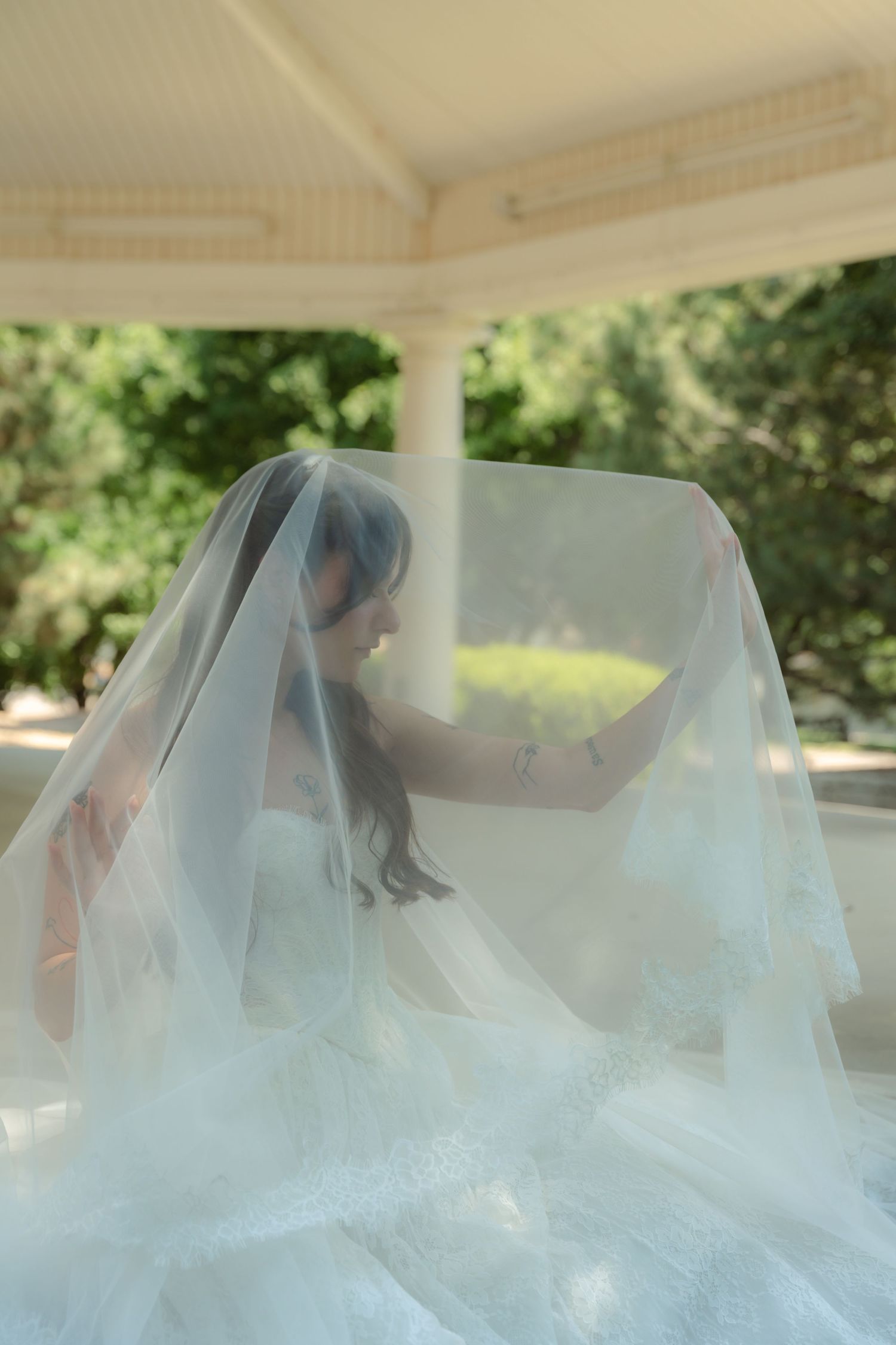 Bride in white wedding gown holding flowing veil in sunlit gazebo surrounded by lush green trees.