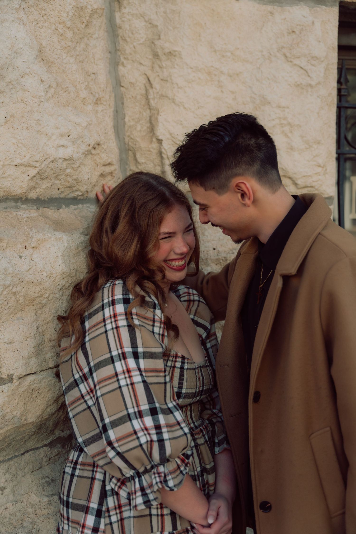 A romantic couple shares a tender moment against a stone wall, with her wearing plaid and him in a brown coat.