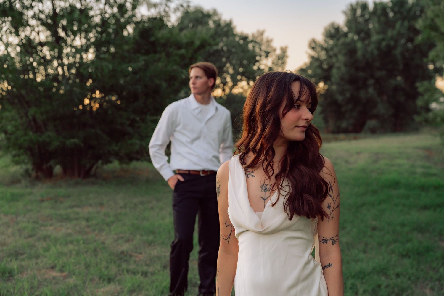 A couple walking through a grassy meadow at dusk wearing white and dark clothing.