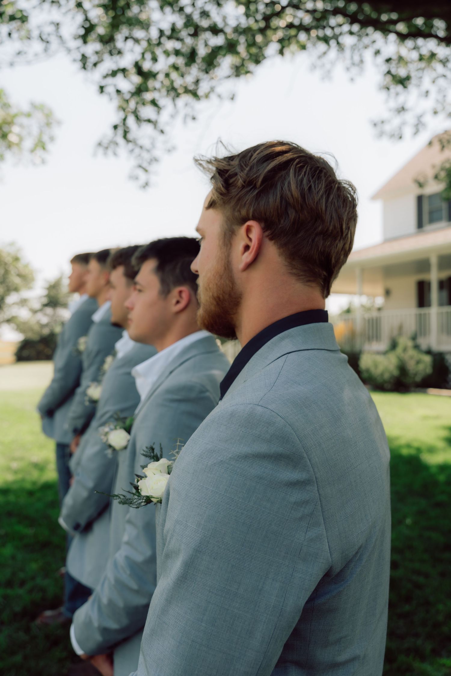 Groomsmen in light gray suits stand in line outdoors near a white house.