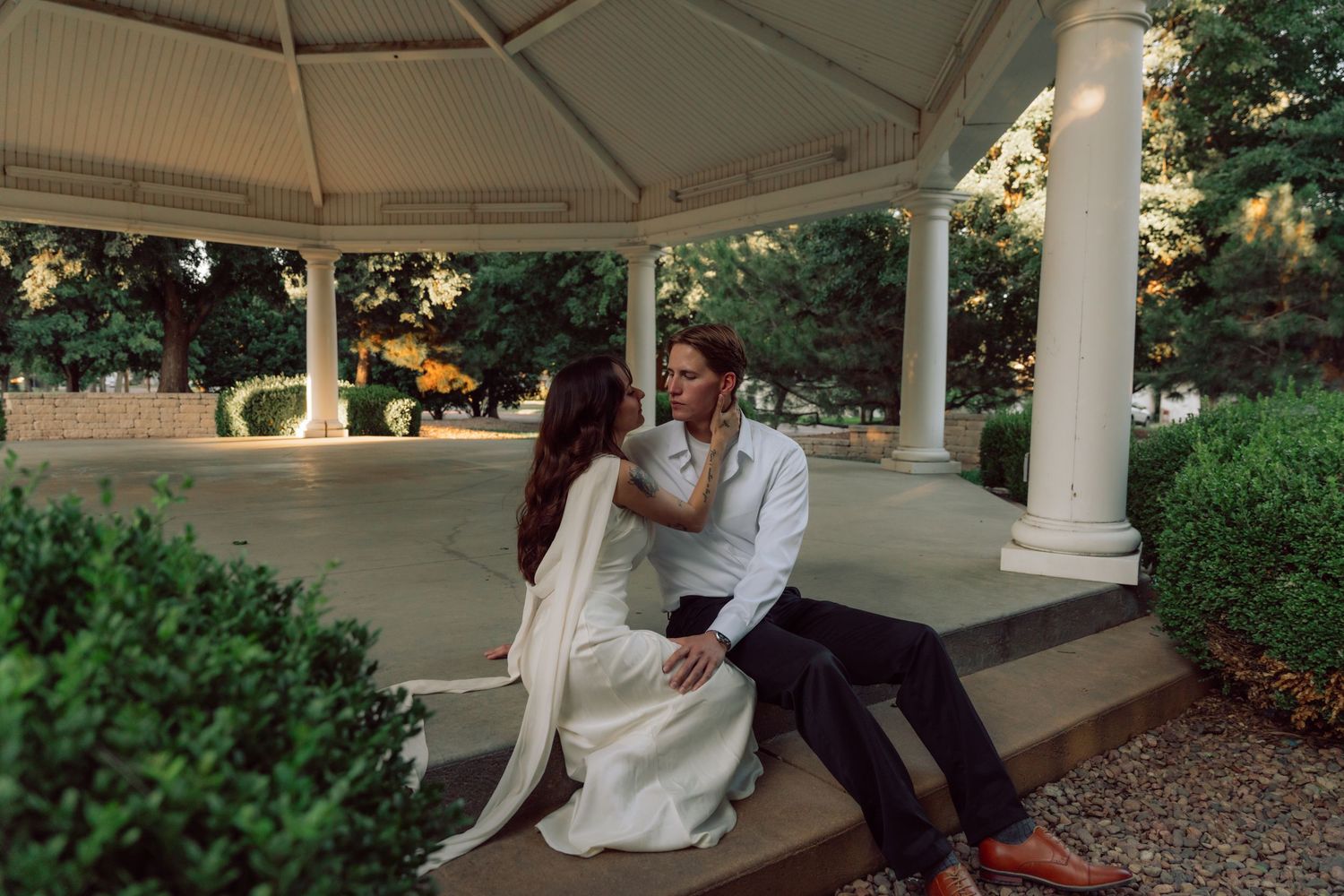 A romantic couple shares an intimate moment in a white gazebo surrounded by lush greenery.