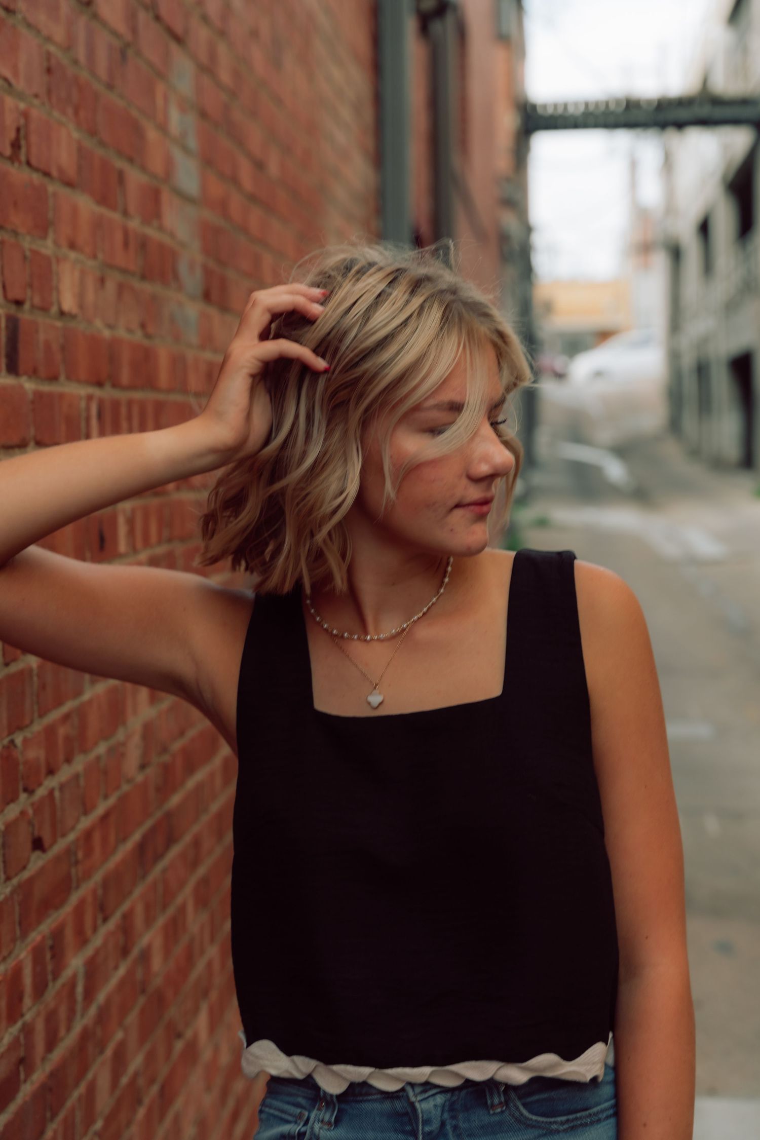 Woman in black sleeveless top poses casually against brick wall in alleyway.