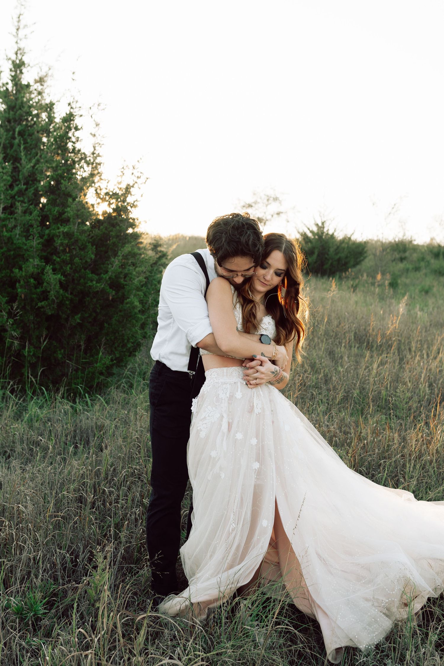 Romantic wedding couple embraces in a sunlit meadow with flowing white dress and natural scenery.
