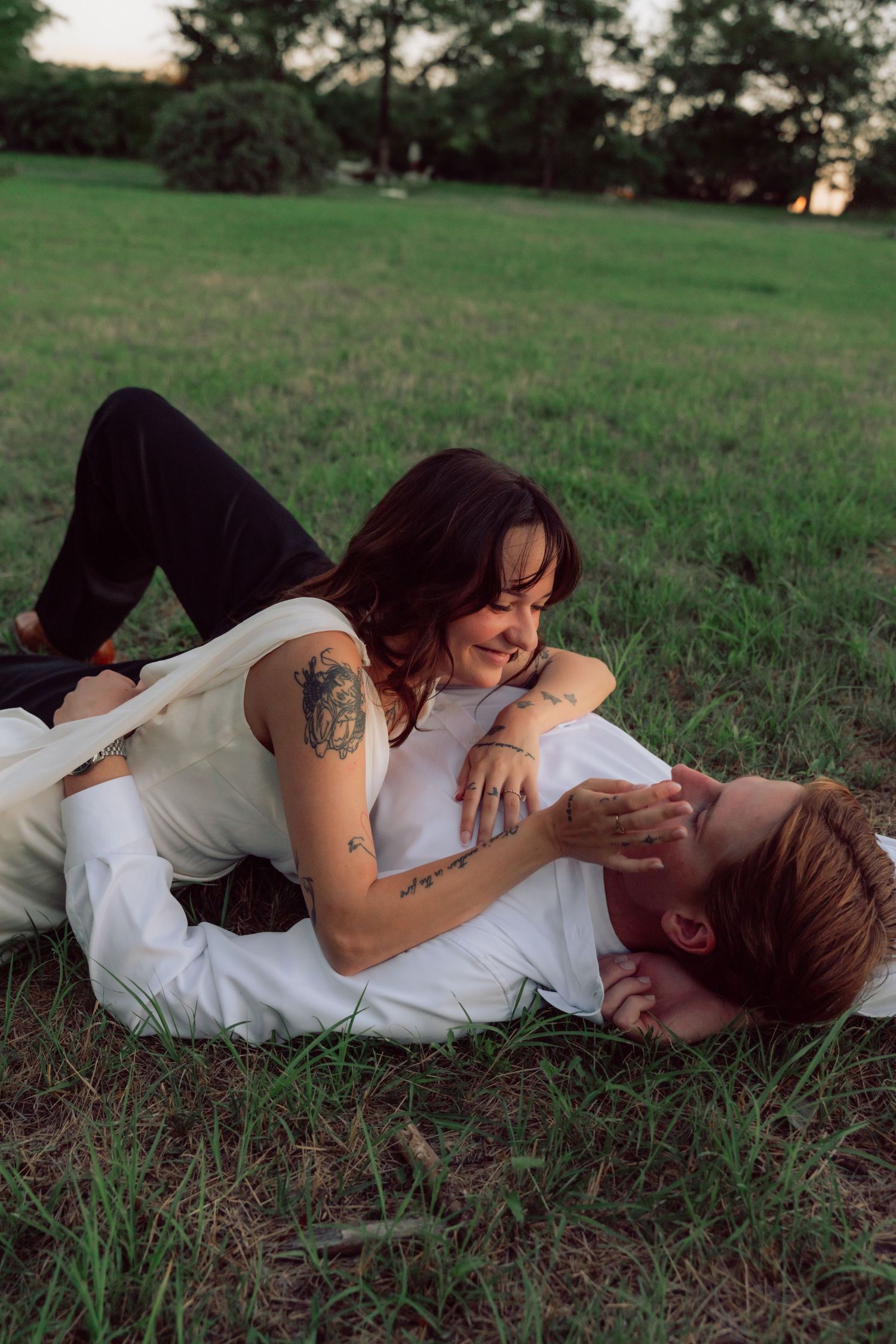 Two people embracing while lying on grass outdoors at dusk.