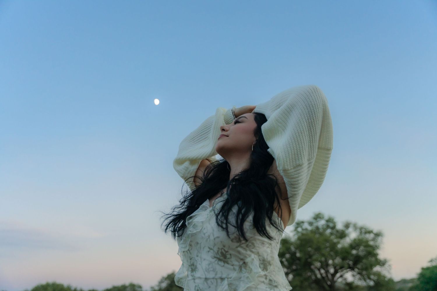 Woman in wide-brim hat gazing at the moon during twilight with trees and blue sky backdrop.