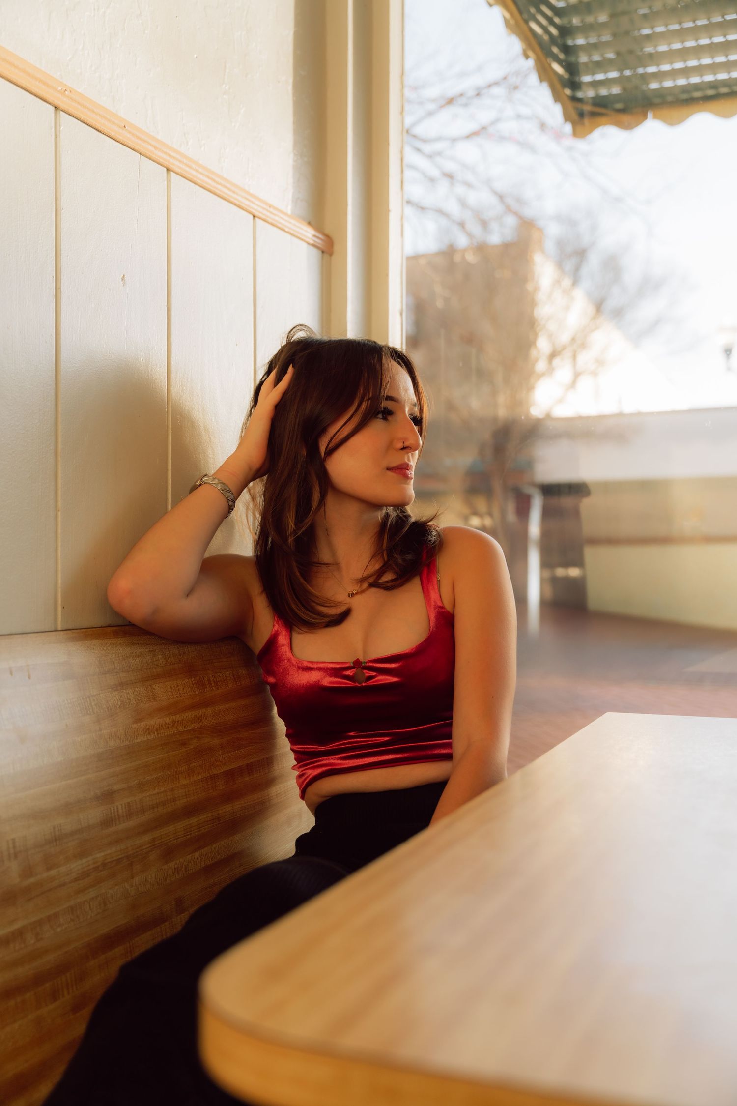 A woman in burgundy attire sits thoughtfully at a wooden table near a window.