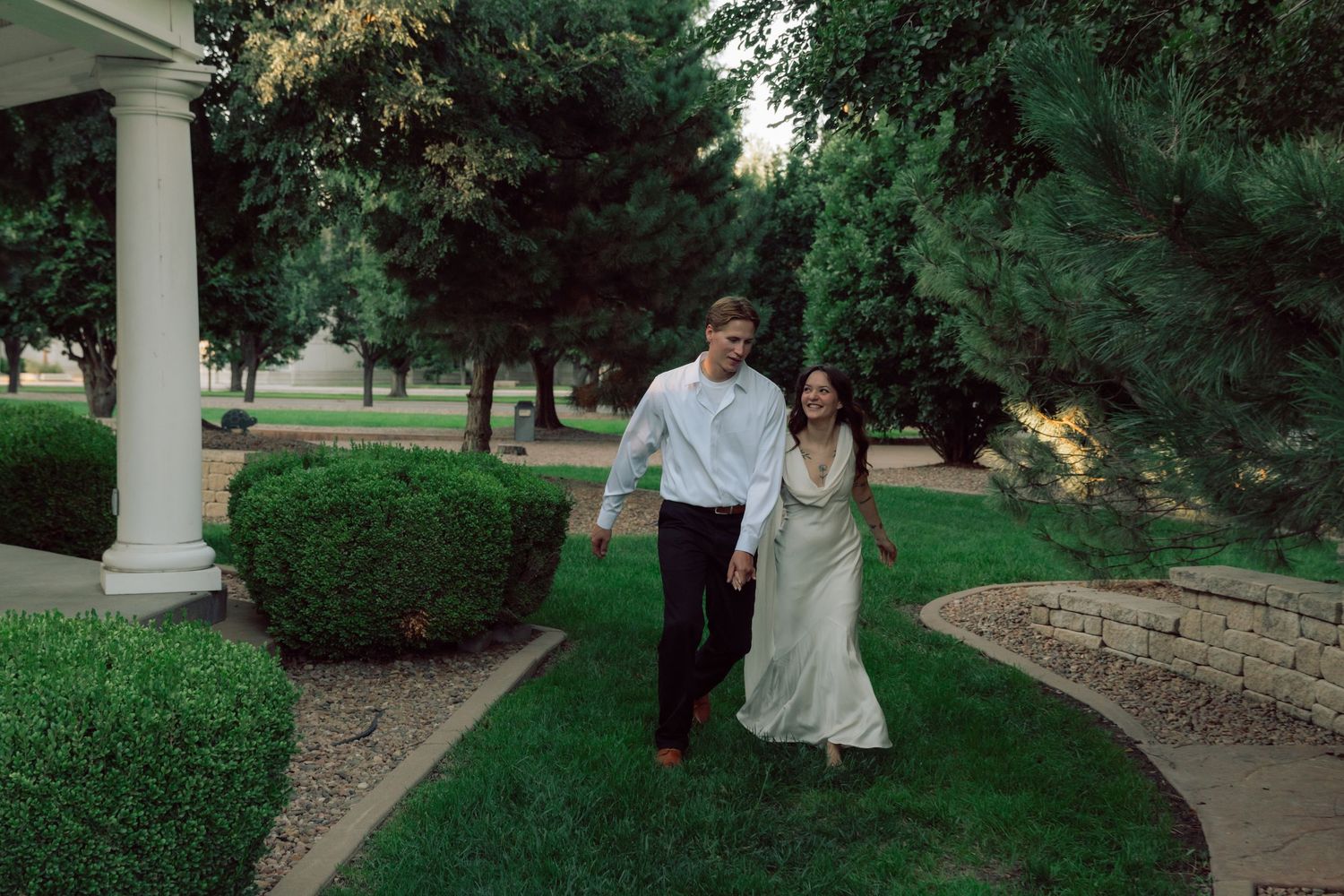 A couple walks together along a columned walkway surrounded by lush greenery.