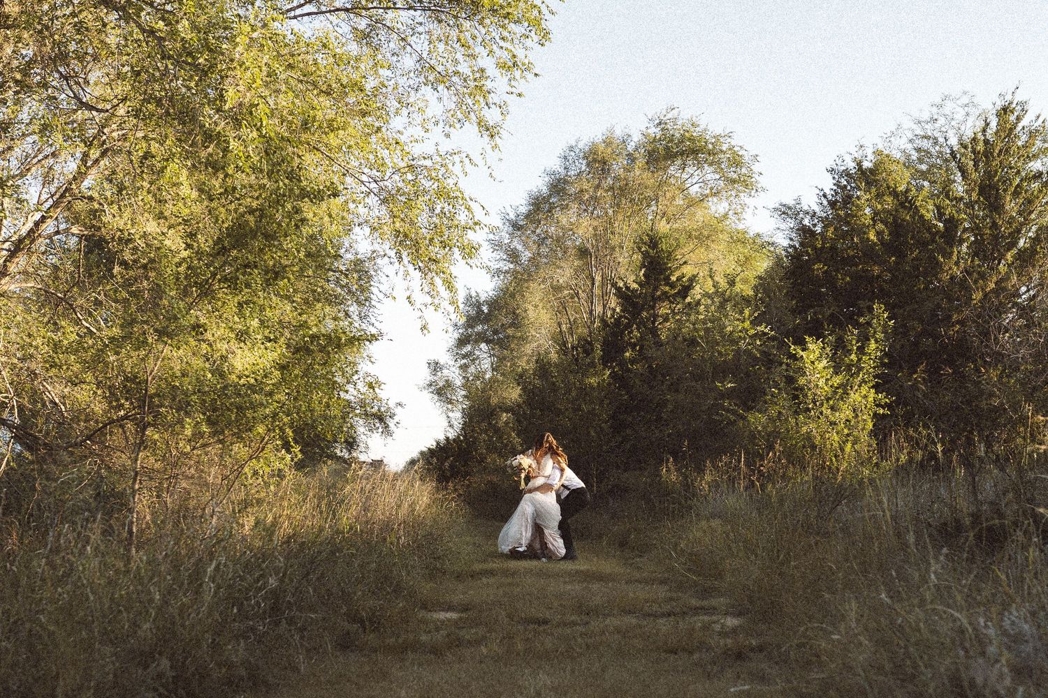 A couple in wedding attire embraces on a grassy hillside during golden hour.