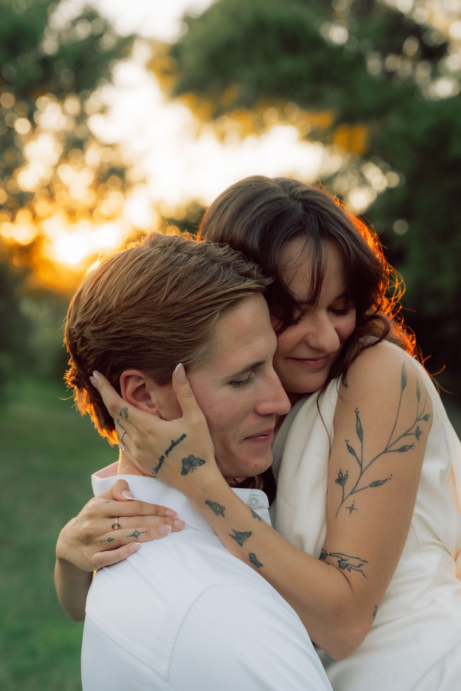 A romantic wedding couple embraces during golden hour sunset with delicate botanical tattoos visible on their arms.