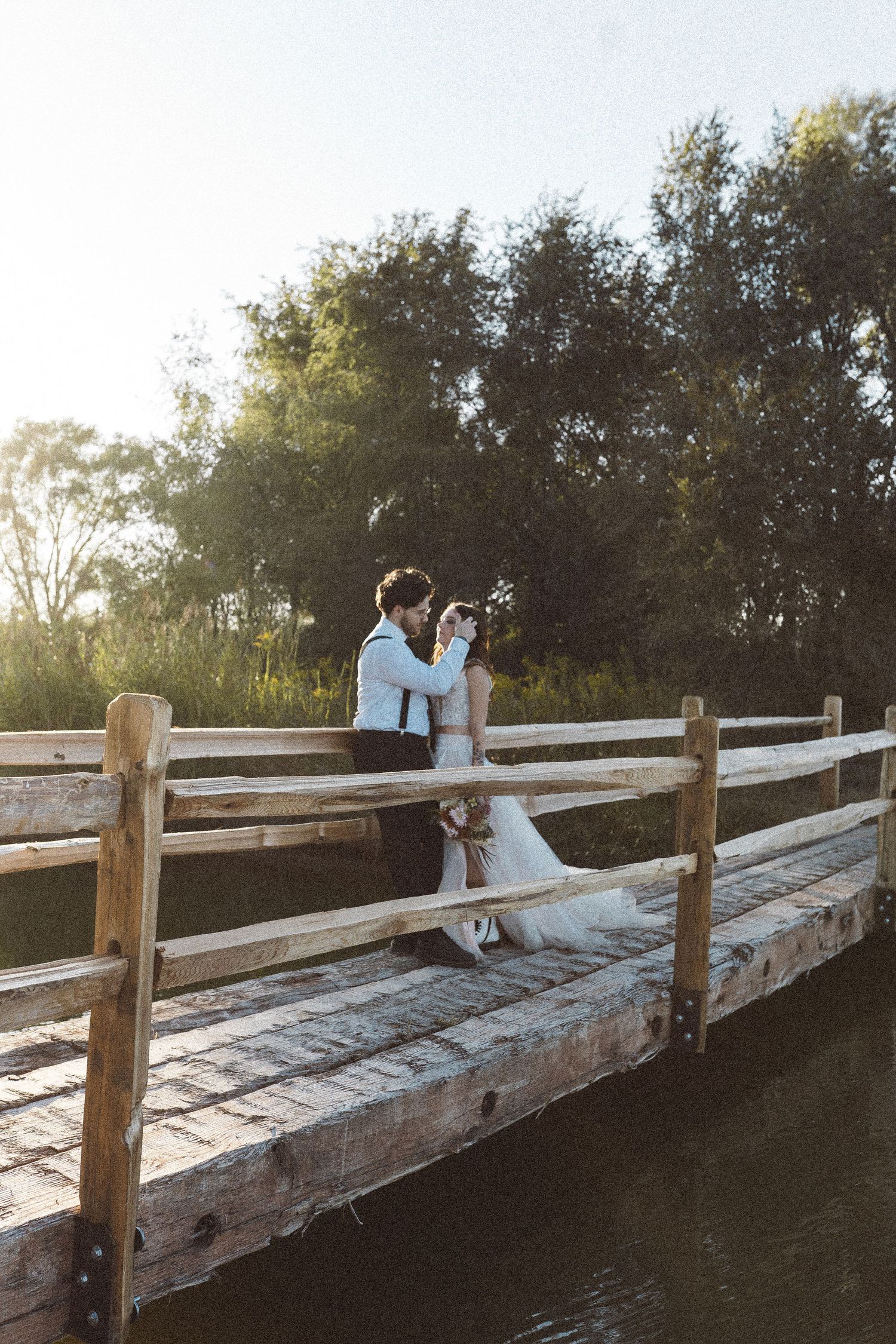 Wedding couple shares intimate moment on rustic wooden bridge over water at sunset.