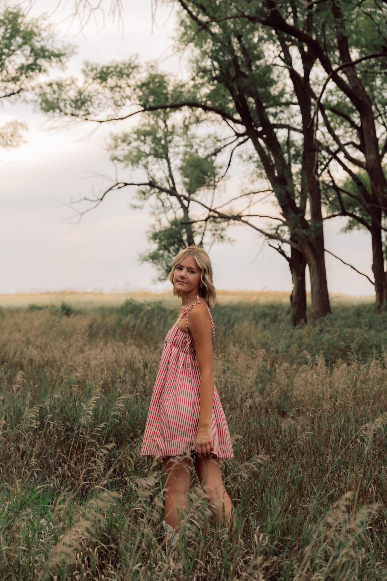 A woman in a pink sundress poses in a grassy field at sunset with tall trees in the background.