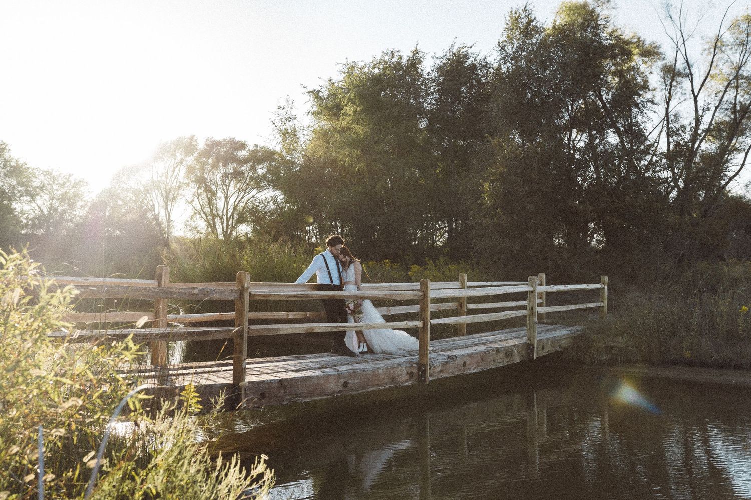 Sunlight filters through trees onto wooden bridge crossing peaceful water.