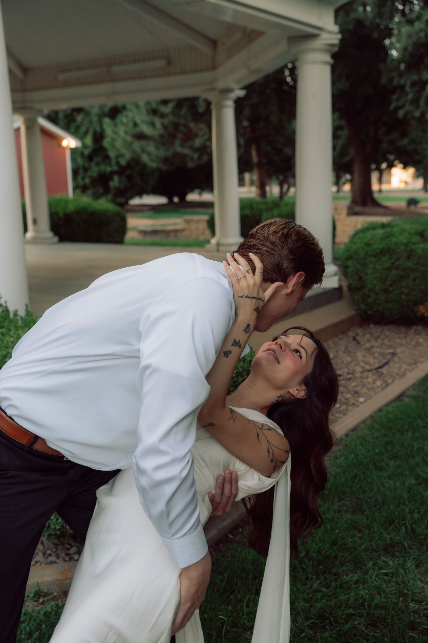 A couple shares an intimate moment near white columns in a garden.