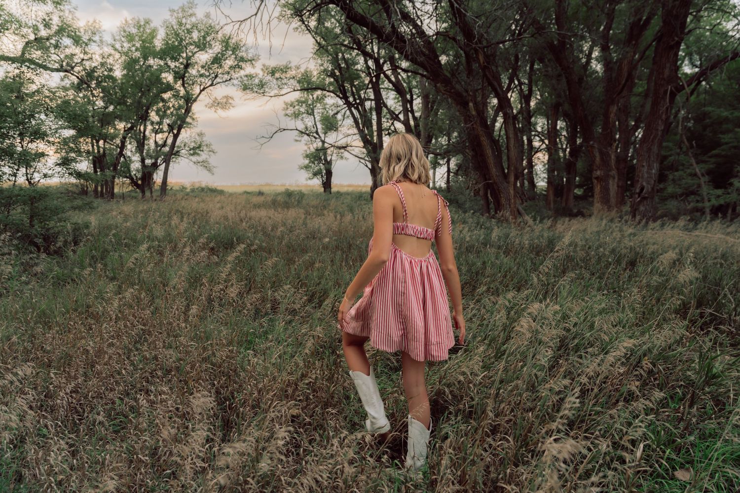 Figure in pink dress stands among trees and tall grass during outdoor photo session.