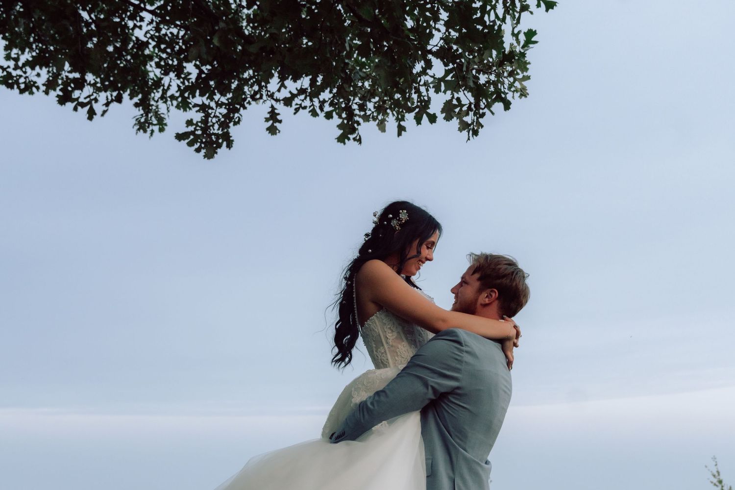 Romantic wedding couple embraces under a tree in an open field during golden hour.