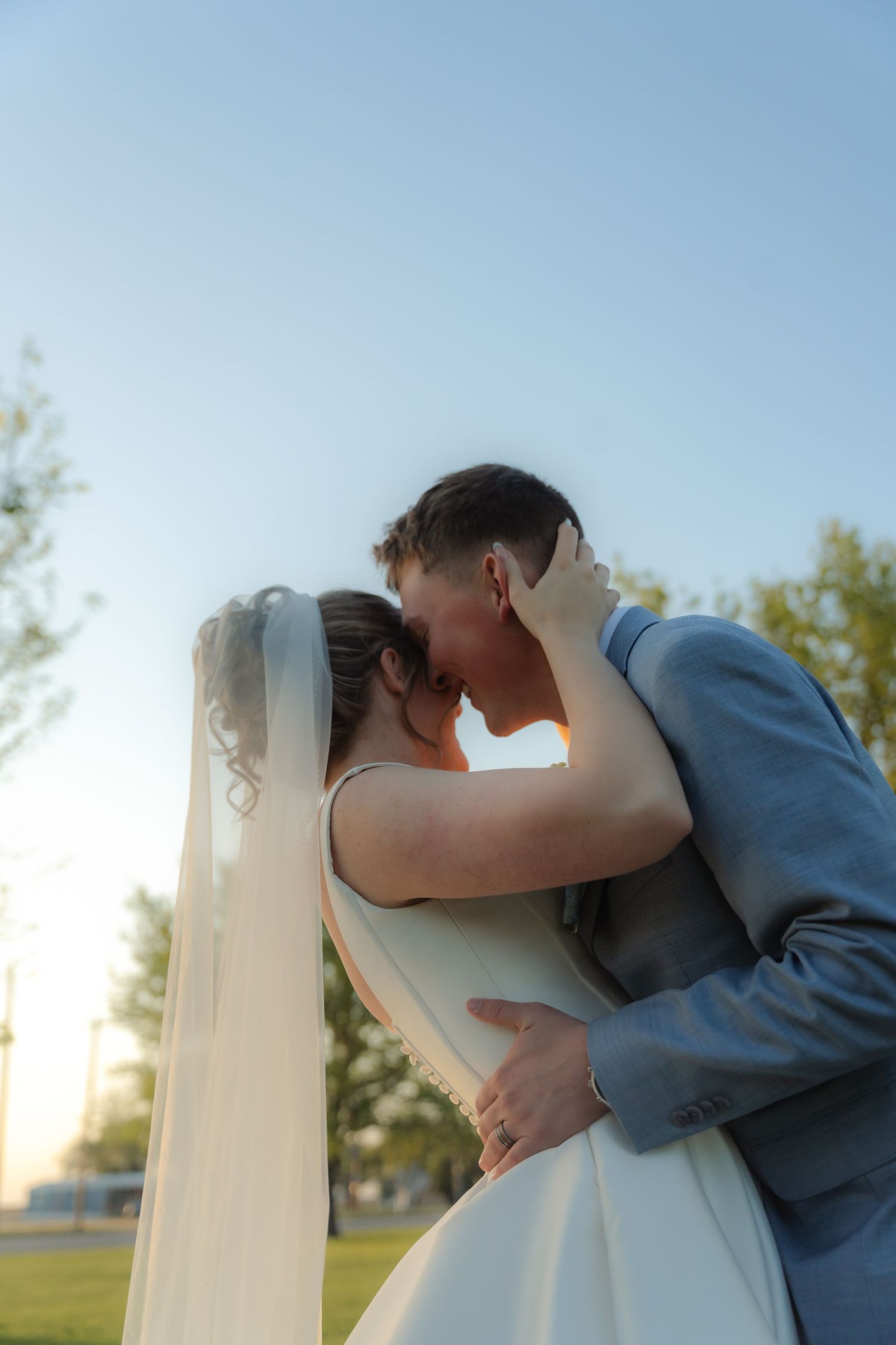 Bride and groom sharing a romantic kiss outdoors at sunset, with wedding veil flowing and blue sky backdrop.