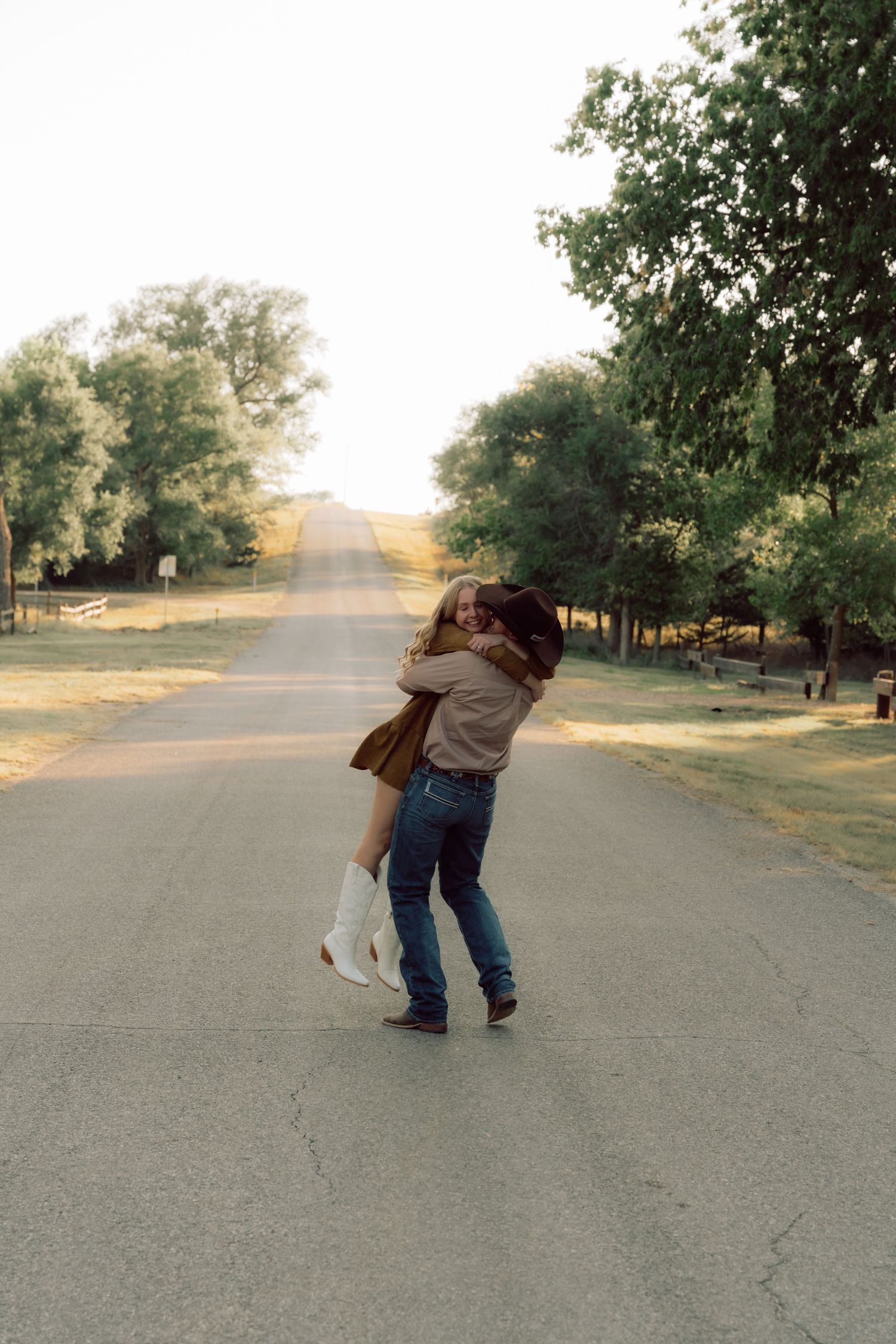 Two people playfully dancing together on an empty road surrounded by trees at sunset.