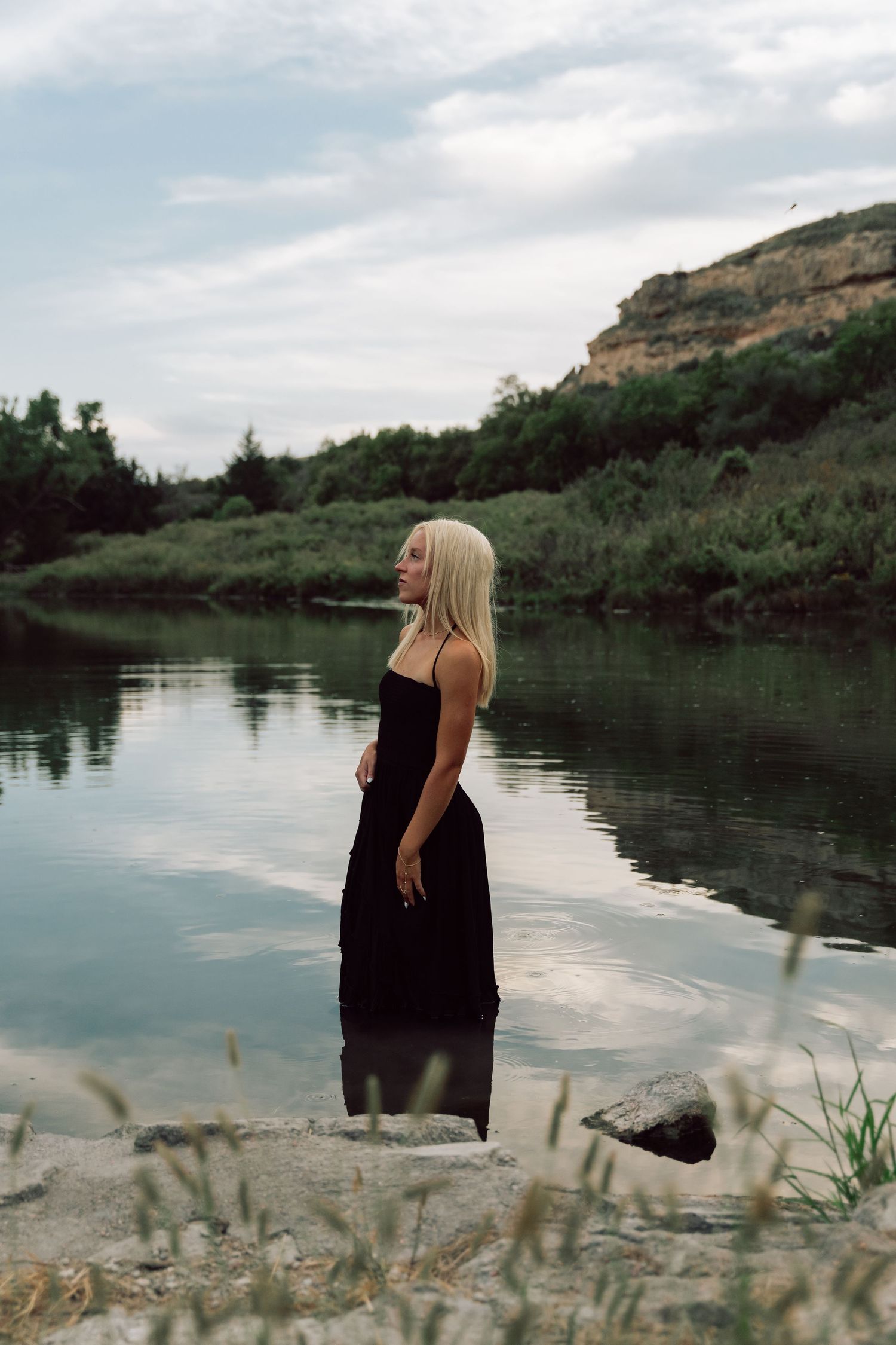 Blonde woman in black maxi dress standing by a calm river with green hills and cloudy sky in background.